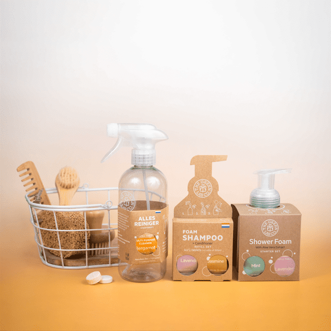 The Green Lab Co. refill products on a warm beige backdrop: a clear “Alles Reiniger” (bergamot) spray bottle, “Foam Shampoo + Conditioner” and “Shower Foam” starter sets, a wire basket with natural brushes and sponge, and two dissolvable tablets.